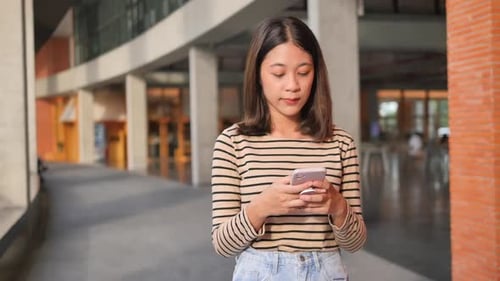 A standing woman succeeds using a smartphone as she walks down the street in front of a building.