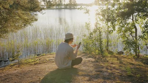 Young Man Surfs Internet on Smartphone Sitting on River Bank