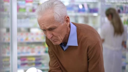 Thoughtful Concentrated Senior Man Choosing Pills in Pharmacy with Blurred Woman at Shelves in