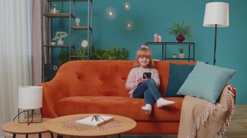 Little Elementary School Kid Girl Using Mobile Phone Technology Device Sitting on Sofa Alone at Home