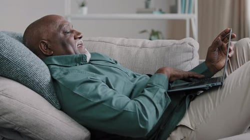 Senior Older Mature African Man Typing on Laptop Lying on Sofa at Home Office Browsing Internet