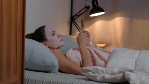 Woman Relaxes in Bed with Face Mask and Phone