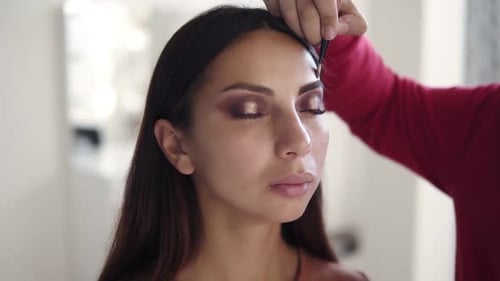 Unerkennbare junge Visagistin, die mit einem schönen jungen Model mit langen braunen Haaren arbeitet