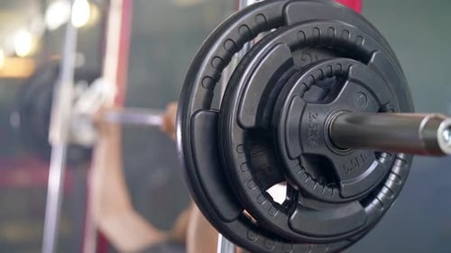 Closeup Shot of Barbell Plates and Bar and Male Athlete Doing Exercises in the Background