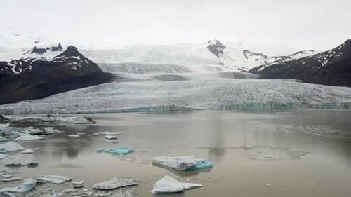 Gletscher in Island mit blauen Eisbrocken im Wasser mit Drohnenvideo, das mit Weitschuss herausgezogen wird.