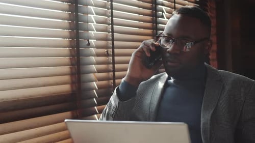 Black Businessman Using Laptop and Talking on Phone in Home Office