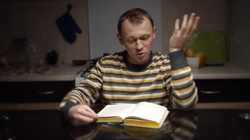 Mann mit Ausdruck liest ein Buch, während er abends in der Küche an einem Tisch sitzt