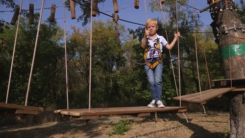 Little Boy with a Safety Carbine Goes on a Rope on the Background of Forest