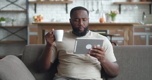 Afrikanischer Mann schaut sich Video auf Tablet-PC an und trinkt Kaffee, während er zu Hause auf dem Sofa sitzt