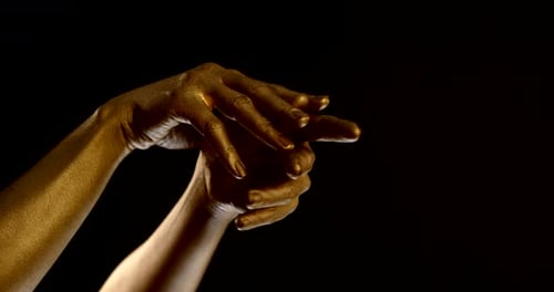 Nahaufnahme weiblicher Hände mit golden leuchtender Haut auf schwarzem Hintergrund. Finger verflechten
sich