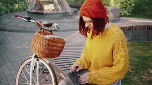 Junge Freelancerin sitzt auf Bank im Stadtpark und arbeitet online am Laptop