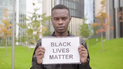 A Young Black Man Shows a Black Lives Matter Sign To the Camera - Office Buildings