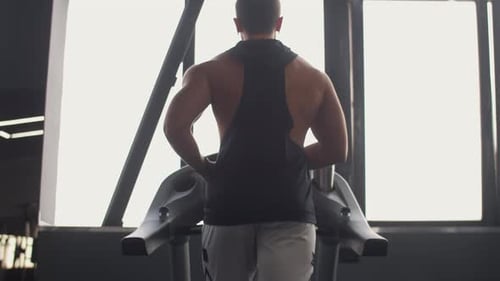 Muscular Man Runs on Treadmill in Gym Warming Up Before Training Back View