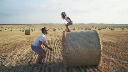 Little Daughter Jumping on Father's Arms From a Big Haycock in Field