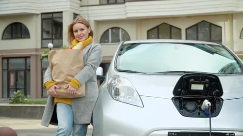 Una mujer se para cerca de su coche eléctrico con la comida y mira a la cámara