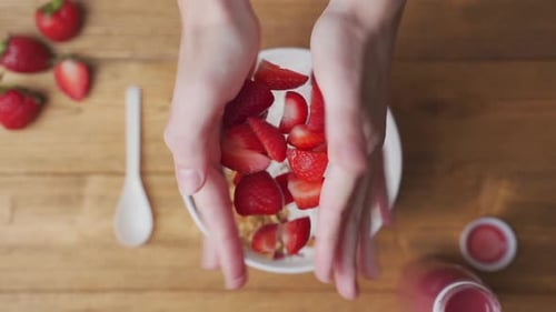 Hands Add Strawberries to Breakfast Bowl with Yogurt
