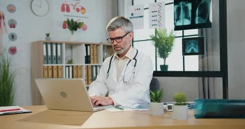 Médecin aux cheveux gris assis sur le lieu de travail dans un cabinet médical et travaillant sur un ordinateur