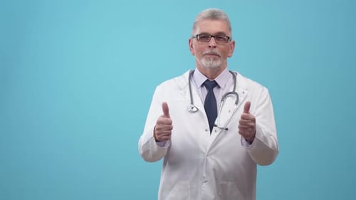 Man Doctor in a Medical Gown with Stethoscope Shows Hands Thumbs Up in the Studio on Blue Background