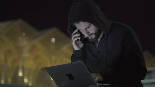 Handsome Hacker in Hood Talking on Phone and Programming on Laptop