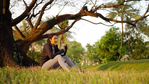 4K Asian woman drinking water from a bottle while jogging at public park in the morning.