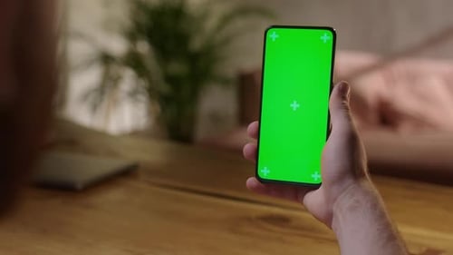Handheld Camera Back View of Young Man at Home Sitting on a Wood Desk Using With Green Mockup Screen