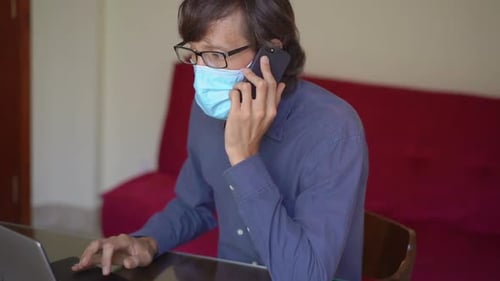 A Young Man Wearing a Face Mask Works From Home During Coronavirus Self-isolation. He Talks on a