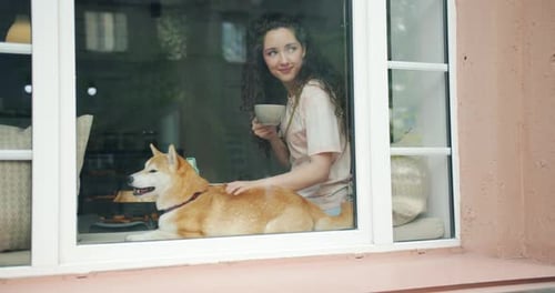 Mujer bonita bebiendo café acariciando lindo cachorro sentado en el alféizar de la ventana