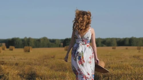 Beautiful Redhaired Woman in a White Dress and Hat Walks Away on the Background of the Field