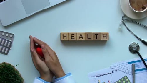 Doctor's Hands Hold Heart on Desk with Health Blocks