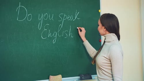 Attractive teacher of English writes on a green board with a chalk.
