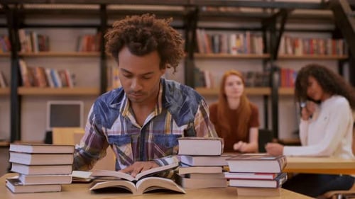 Dark Skinned Young Male Sitting at Table in Library Surrounded By Books Making Notes While Two