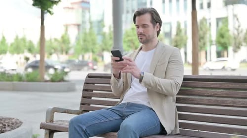 Man Typing on Phone on Bench in City