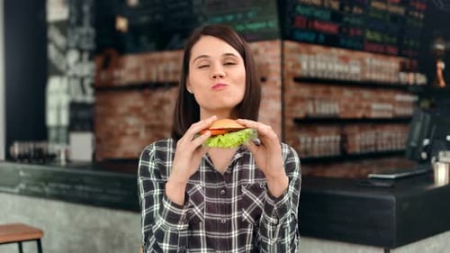 Happy Casual European Female Eating Appetizing Juicy Burger at Fast Food Restaurant Medium Shot