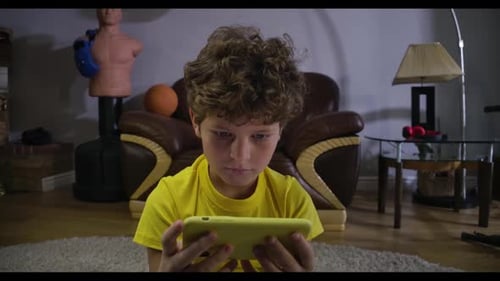 Close-up Portrait of Charming Curly-haired Boy Looking at Smartphone Screen and Smiling. Caucasian