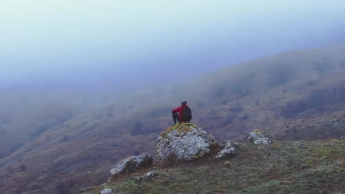 Un viajero con una chaqueta roja se sienta sobre una piedra sobre un fondo de montañas