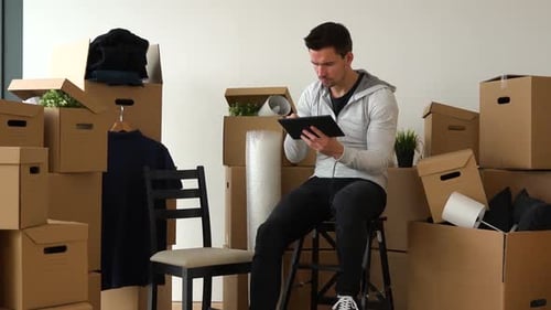 A Moving Man Sits on a Chair in an Empty Apartment and Works on a Tablet, Then Smiles at the Camera