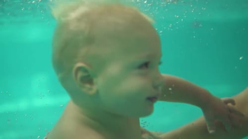 Cute Blonde Toddler is Swimming Under the Water in the Swimming Pool Until His Mother Helps Him to
