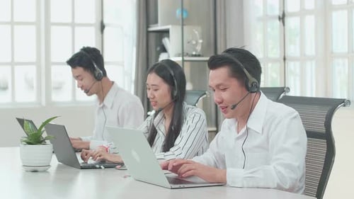 Three Asian Call Centre Agents Wearing Headsets Speaking To Customers At The Office