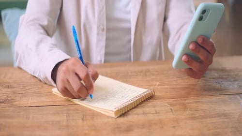 Closeup Man Hands Making Notes in Notebook and Holding Mobile Phone at Table