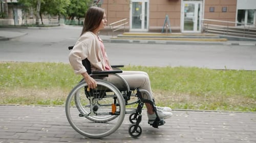 Side View of Beautiful Disabled Woman Moving in Wheelchair Along City Street