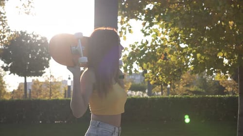 Woman Carries Skateboard Through Park in Sunny Weather
