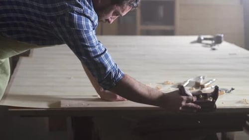 Male Carpenter in Shirt in Dark Workshop Planing Wood