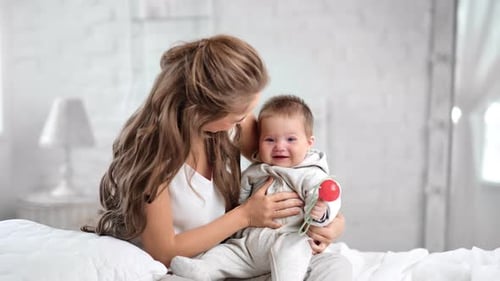 Caring Beautiful Mother Swaying Smiling Cute Baby Having Positive Emotion at Morning White Bedroom