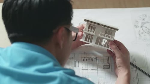 Close Up Of A Man Engineer Looking At Paper Model Of House At The Office, Over Shoulder