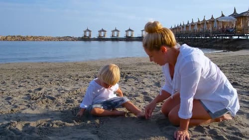 Little Boy in a White Shirt Plays in the Sand on the Beach with Mom