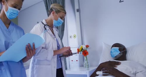 Diverse female doctors talking to male patient in hospital bed all wearing face masks
