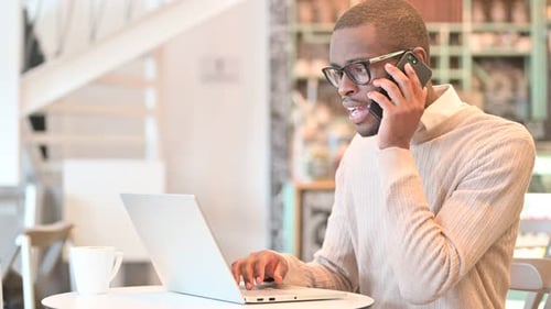 African Man with Laptop Talking on Smartphone in Cafe