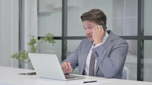 Middle Aged Man Talking on Phone while using Laptop in Office