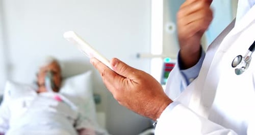 Doctor Checks Tablet Near Elderly Patient in Hospital Bed