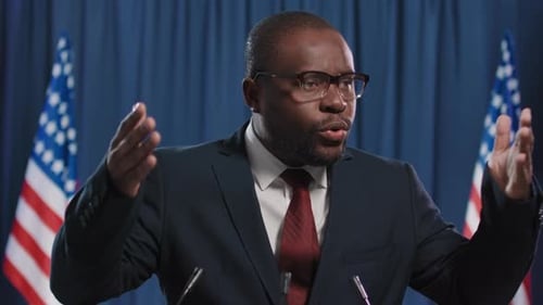 Adult Male Giving Speech at Podium with Flags